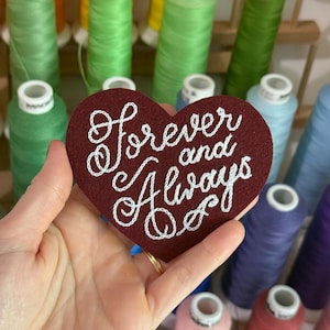 May include: A heart-shaped dark red felt patch featuring the white embroidered text "forever and Always" in a cursive script. The patch is held against a background of various colorful sewing thread spools arranged on a wooden rack.