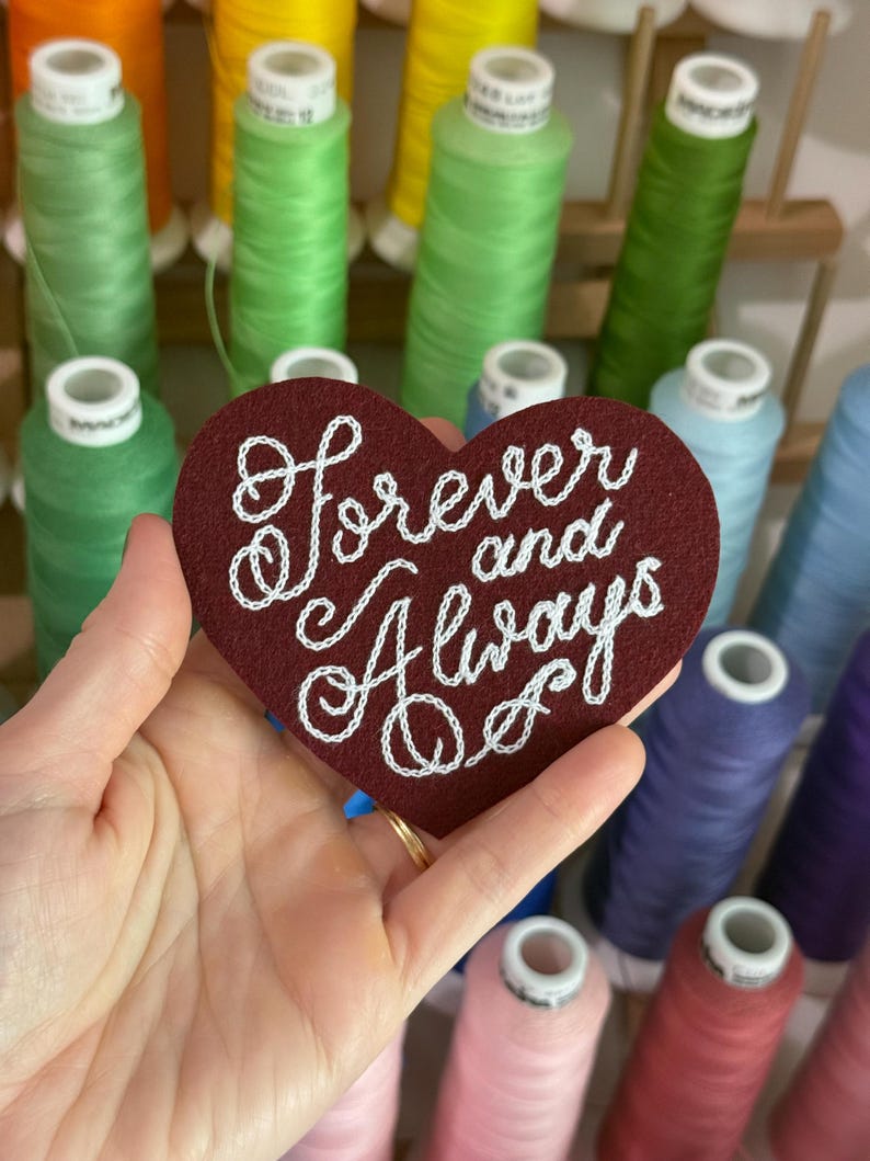 May include: A heart-shaped dark red felt patch featuring the white embroidered text "forever and Always" in a cursive script. The patch is held against a background of various colorful sewing thread spools arranged on a wooden rack.