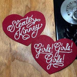 May include: Two heart-shaped red felt patches featuring white embroidered text. One patch displays the words Sleater Kinney in a cursive script, while the other patch features the repeated text Girls! Girls! Girls! in a matching white cursive style.