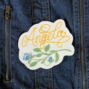 K&ouml;nnte beinhalten: Gestickter Aufn&auml;her mit dem Namen "Angela" in gelber Schrift, begleitet von gr&uuml;nen Bl&auml;ttern und blauen Blumen. Der Aufn&auml;her hat einen wei&szlig;en Hintergrund mit einem gewellten Rand. Der Hintergrund ist eine blaue Jeansjacke.