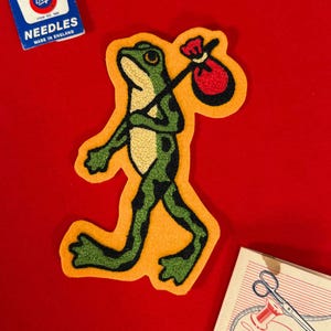 Peut inclure: Écusson en feutre représentant une grenouille verte à taches noires, portant un sac rouge sur un bâton. La grenouille marche sur un fond jaune. L'écusson est sur une surface rouge. L'image comprend également une boîte d'aiguilles et un kit de couture.