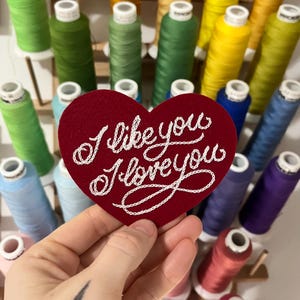 hand holding red custom felt heart patch that says "I like you I love you" in fancy cursive script