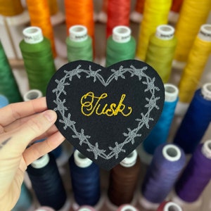 Puede incluir: Un parche de fieltro negro en forma de corazón con la palabra "Tusk" bordada en hilo dorado. El corazón está delineado con bordados de alambre de púas gris. En el fondo, hay carretes de hilo coloridos.