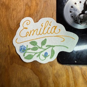 K&ouml;nnte beinhalten: Gestickter Aufn&auml;her in Wolkenform mit dem Namen "Emilia" in goldfarbenem Faden. Unter dem Namen ist ein Blumendesign mit blauen Bl&uuml;ten und gr&uuml;nen Bl&auml;ttern aufgestickt. Der Aufn&auml;her liegt auf einer Holzoberfl&auml;che.