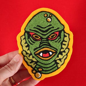 May include: Embroidered patch featuring a green creature face with red eyes, a white mouth, and yellow accents. The patch has a yellow border and is set against a red background. The design is detailed and stylized, suitable for apparel or accessories.