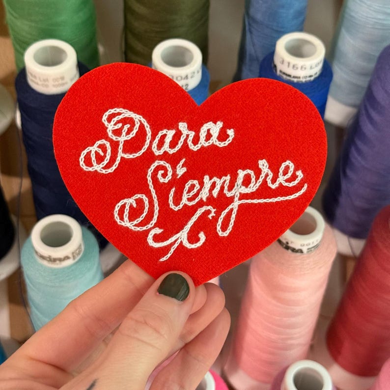May include: A red heart-shaped felt patch featuring the white embroidered text Para Siempre in a cursive script. The patch is positioned in front of an assortment of colorful sewing thread spools in various shades of blue, green, pink, and red.