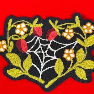 Pode incluir: Patch bordado com fundo preto, com uma teia de aranha branca, bagas vermelhas e flores e folhas amarelas. O patch está em um fundo vermelho.