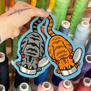 May include: Two embroidered patches featuring cats with wings, one in gray with black stripes and one in orange with brown stripes. Both cats have green eyes, red accents, and are positioned above a gray bowl on a light blue felt backing.