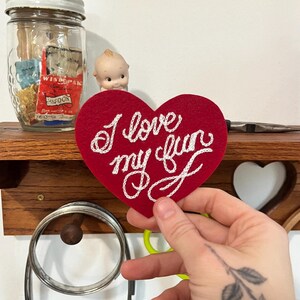 hand holding red custom felt heart patch that says "I love my fun" in fancy cursive script