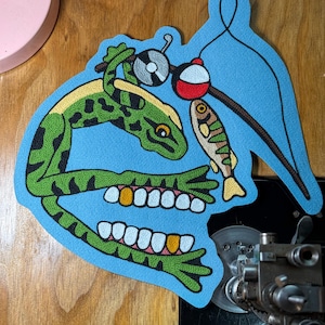 May include: A light blue embroidered patch featuring a green frog with black spots and human-like teeth, including gold teeth. The frog holds a fishing rod with a red and white bobber and a small fish attached to the line, all on a blue background.