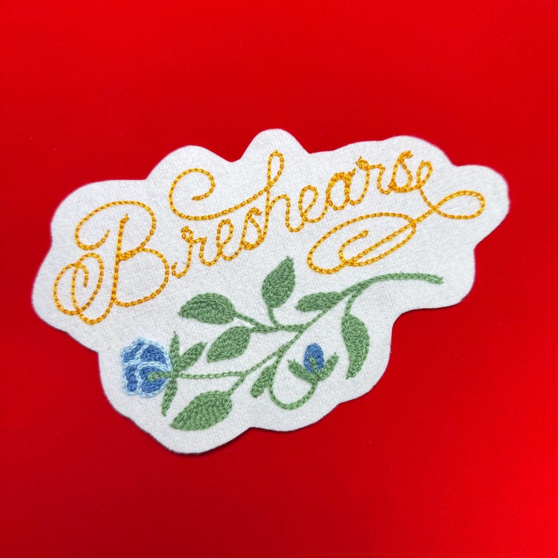 K&ouml;nnte beinhalten: Gestickter Aufn&auml;her mit dem Wort "Breshearse" in goldener Schrift. Der Aufn&auml;her zeigt ein florales Design mit gr&uuml;nen Bl&auml;ttern und blauen Blumen auf wei&szlig;em Grund. Der Aufn&auml;her hat einen gewellten Rand und ist vor rotem Hintergrund platziert.