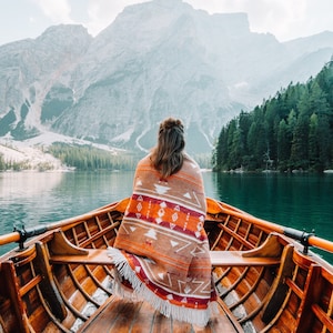 Könnte beinhalten: Eine Person sitzt in einem Holzboot auf einem See und trägt eine bunte, gemusterte Decke. Der See ist von Bergen und Bäumen umgeben.