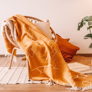 Könnte beinhalten: Ein geflochtener Korbstuhl mit einer senfgelben Wolldecke darüber. Die Decke hat ein weißes geometrisches Muster. Auf dem Stuhl liegen zwei Kissen, eines cremefarben und eines orange.
