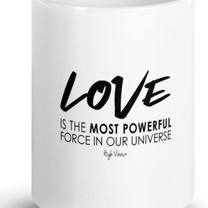 Pode incluir: Caneca de café de cerâmica branca com texto preto que diz "LOVE IS THE MOST POWERFUL FORCE IN OUR UNIVERSE" e "High Vibes" em texto menor.