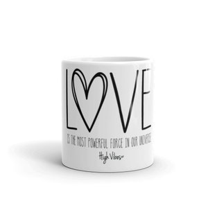 Pode incluir: Caneca de cerâmica branca com texto preto que diz "LOVE is the most powerful force in our universe" e um gráfico de coração preto. O texto "High Vibes" também está impresso em preto na parte inferior da caneca.