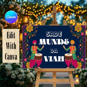 May include: A rectangular sign on a wooden easel featuring a dark blue background with a floral border. The text reads SADE MUNDE DA VIAH. Two illustrated figures in traditional attire are playing dhol drums on either side of the text.