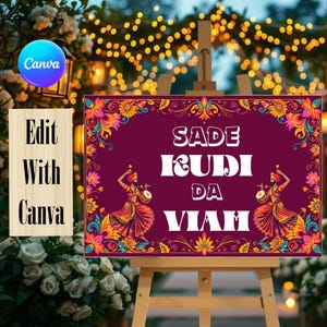 Puede incluir: Un letrero rectangular con fondo granate oscuro y un borde floral decorativo en naranja, amarillo y rosa. El texto SADE KUDI DA VIAH aparece en blanco, flanqueado por dos ilustraciones de bailarines con vestimenta tradicional y tambores.