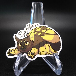 Puede incluir: Una pegatina con un gato de dibujos animados con pelaje amarillo y marrón. El gato está enroscado con las patas metidas debajo de su cuerpo. La pegatina tiene la palabra "Spittern" escrita en cursiva sobre el gato.