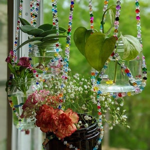 May include: Four glass jars with plants hanging from a window using colorful beaded strings. The jars are filled with water and contain a variety of plants, including a succulent, a vine, and a bouquet of flowers.