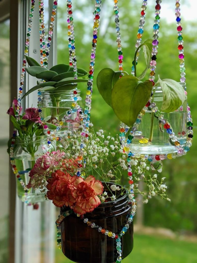 May include: Four glass jars with plants hanging from a window using colorful beaded strings. The jars are filled with water and contain a variety of plants, including a succulent, a vine, and a bouquet of flowers.