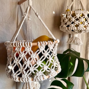 May include: Two macrame hanging baskets with wooden dowels and tassels. The baskets are filled with fruit, including yellow bananas, green apples, and pears. The baskets are hanging against a beige background.