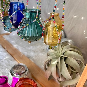 May include: A set of four glass vases hanging from colorful beaded macrame plant hangers. The vases are in various shapes and sizes, including a blue vase, a green vase, a clear vase, and a yellow vase. The vases are hanging from a wooden shelf with a green air plant on the shelf.