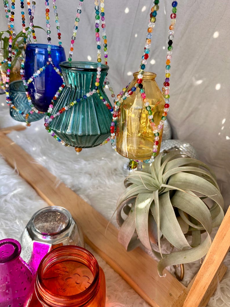 May include: A set of four glass vases hanging from colorful beaded macrame plant hangers. The vases are in various shapes and sizes, including a blue vase, a green vase, a clear vase, and a yellow vase. The vases are hanging from a wooden shelf with a green air plant on the shelf.
