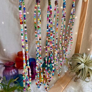 May include: A display of colorful beaded necklaces with various charms, including a small eye charm, hanging from a wooden stand.