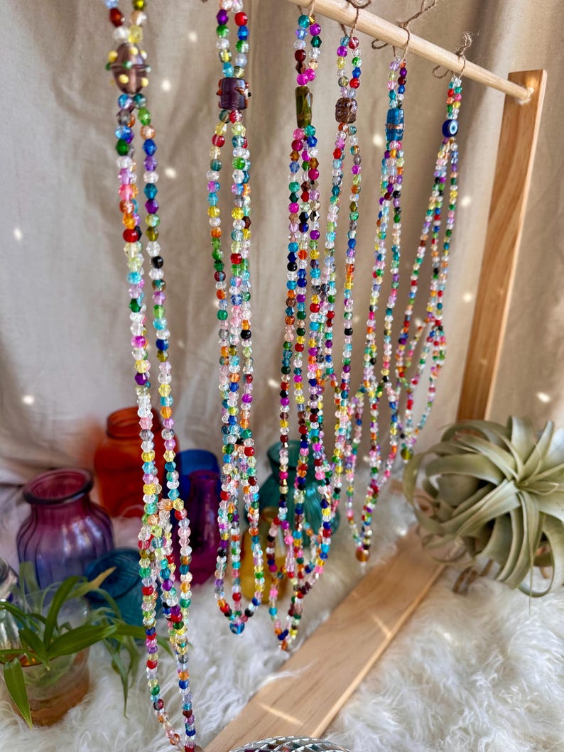 May include: A display of colorful beaded necklaces with various charms, including a small eye charm, hanging from a wooden stand.