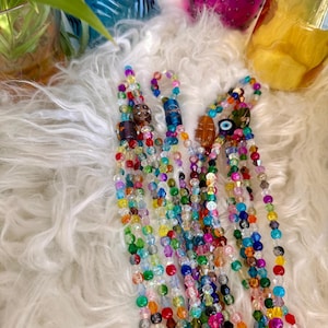 May include: A collection of colorful beaded necklaces with various charms, including an evil eye charm, a brown and gold charm, and a blue and gold charm. The necklaces are laid out on a white fluffy surface.