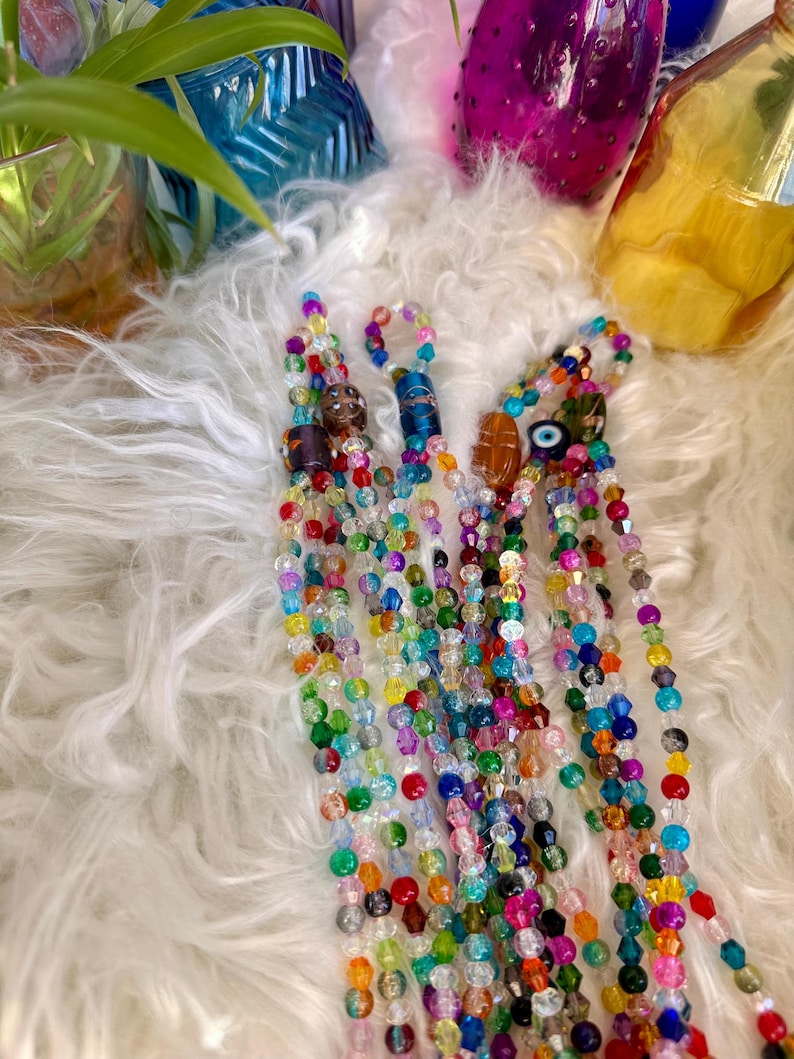 May include: A collection of colorful beaded necklaces with various charms, including an evil eye charm, a brown and gold charm, and a blue and gold charm. The necklaces are laid out on a white fluffy surface.