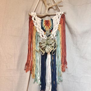 May include: A macrame wall hanging featuring a natural antler branch and colorful yarn. The design incorporates shades of orange, yellow, blue, and white, with a central air plant. The piece is a bohemian-style home decor item.