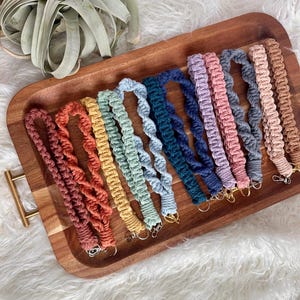 May include: A collection of 12 colorful crocheted lanyards with gold keychains. The lanyards are arranged in a rainbow pattern on a wooden tray.