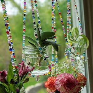May include: Three glass jars with plants hanging from colorful beaded chains. The jars are filled with water and the plants are growing roots. The jars are hanging in front of a window with green foliage visible outside.