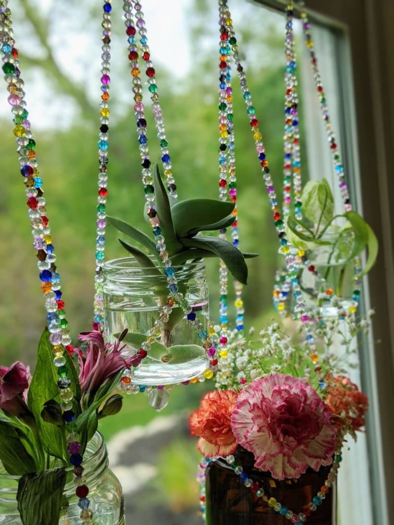May include: Three glass jars with plants hanging from colorful beaded chains. The jars are filled with water and the plants are growing roots. The jars are hanging in front of a window with green foliage visible outside.