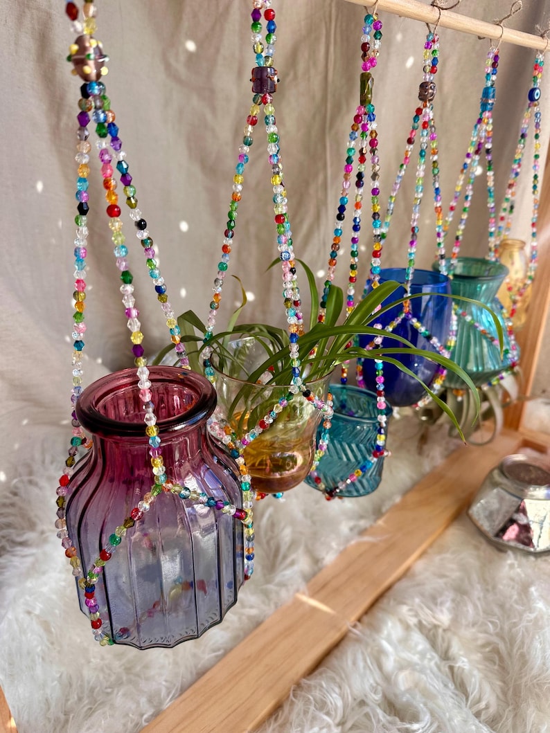 May include: Five colorful glass vases hang from a wooden rack, suspended by colorful beaded macrame plant hangers. The vases are filled with plants and greenery.