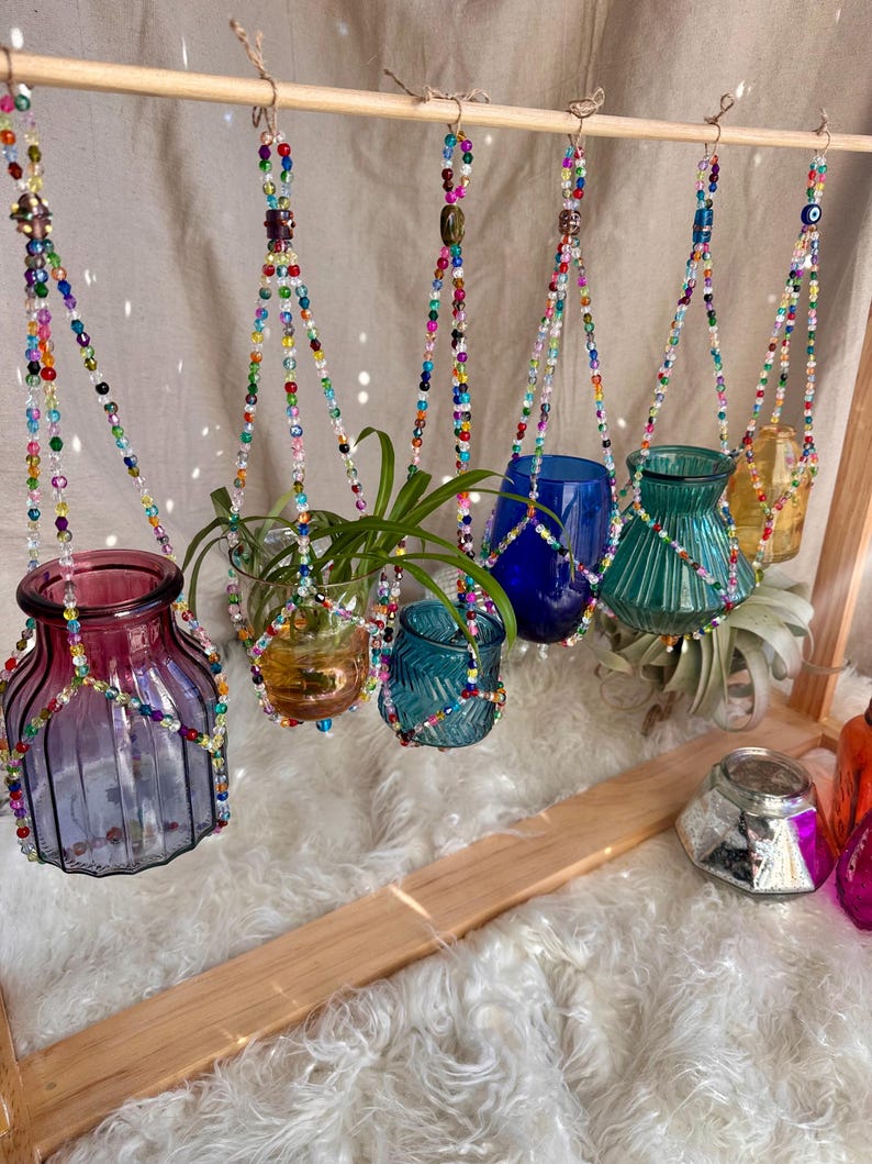 May include: A set of six colorful glass vases hanging from beaded macrame plant hangers. The vases are in various colors, including blue, green, purple, and yellow. The hangers are made of small, colorful beads and are strung in a geometric pattern.