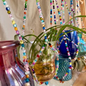 May include: A set of five glass vases with colorful beaded hangers. The vases are filled with water and plants. The vases are hanging from a wooden dowel rod.