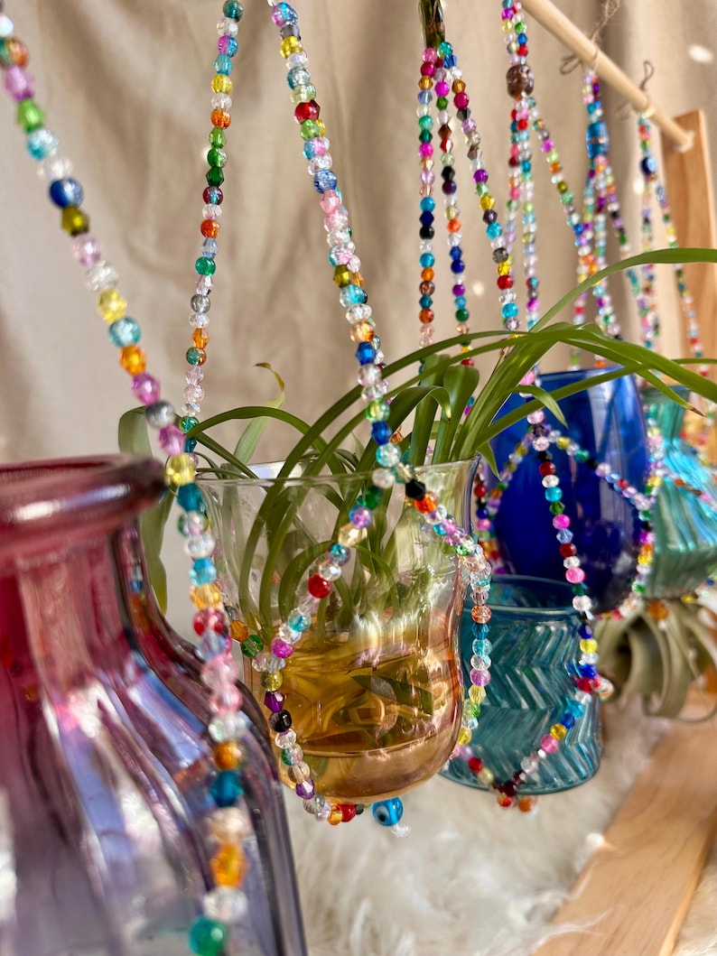 May include: A set of five glass vases with colorful beaded hangers. The vases are filled with water and plants. The vases are hanging from a wooden dowel rod.