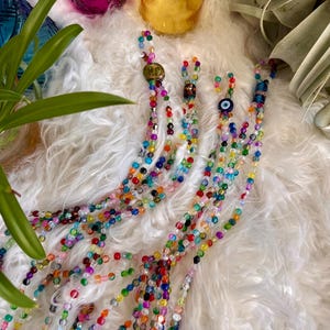 May include: Multicolored beaded necklace with an evil eye charm. The necklace is made of small, round beads in various colors, including blue, green, red, and yellow. The necklace is lying on a white fluffy surface.