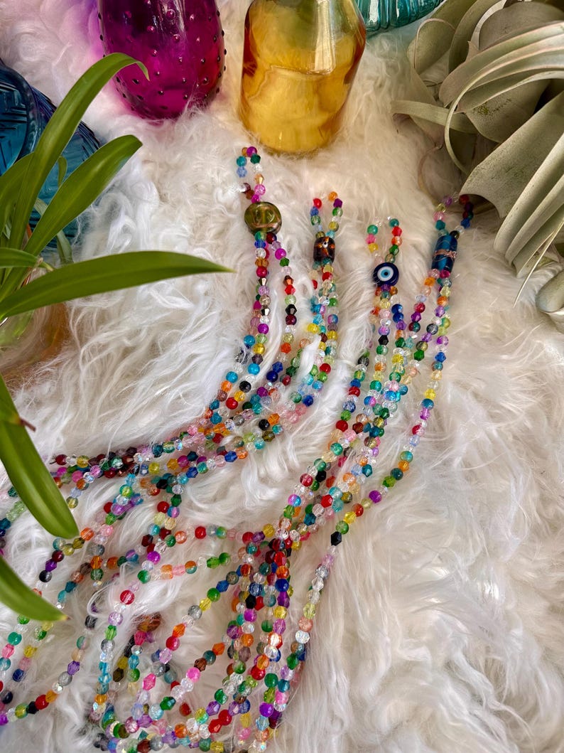 May include: Multicolored beaded necklace with an evil eye charm. The necklace is made of small, round beads in various colors, including blue, green, red, and yellow. The necklace is lying on a white fluffy surface.