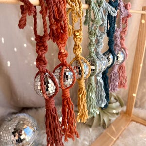 May include: A set of seven colorful macrame tassel ornaments with disco ball accents. Each tassel is made with a different color of yarn, including red, orange, yellow, green, blue, purple, and pink. The tassels are tied to a wooden dowel rod.