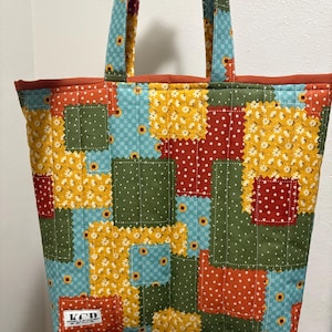 以下が含まれることがあります： パッチワークデザインのキルティングトートバッグ。青、緑、黄、赤の生地に花柄と水玉模様の正方形が特徴です。ハンドルと上部のトリムは無地のオレンジ色です。バッグには「KCD」と書かれたラベルが付いています。