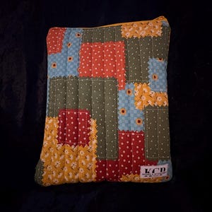 Puede incluir: Una bolsa acolchada de patchwork con cremallera amarilla. La tela presenta cuadrados rojos, azules, amarillos y verdes con estampados florales y de lunares. La etiqueta dice "KCD Apparel & Accessories."