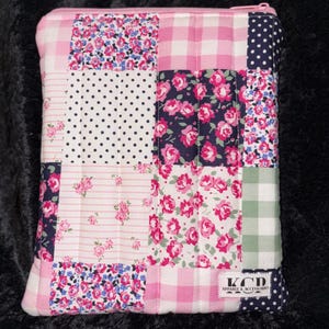 Puede incluir: Un estuche con cremallera de patchwork con una variedad de estampados florales, de lunares y de cuadros en rosa, blanco y azul marino. El estuche tiene una cremallera rosa y una etiqueta que dice "KCD Apparel & Accessories".