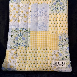 May include: A quilted patchwork pillow cover featuring a variety of floral and patterned fabrics in yellow, white, and blue. The design includes small yellow flowers, blue floral sprigs, and a yellow gingham pattern. The label reads "KCD Apparel & Accessories."