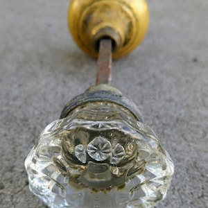 May include: A vintage door knob set featuring a clear, faceted glass knob on one end and a smooth, rounded brass knob on the other, connected by a square metal spindle. The glass knob displays a starburst pattern and shows signs of age.