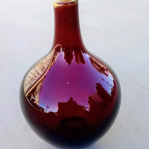 May include: A deep burgundy glass vase with a spherical body and a narrow, cylindrical neck. The surface is highly reflective, showing distorted environmental reflections across its smooth, glossy, and dark red exterior finish.