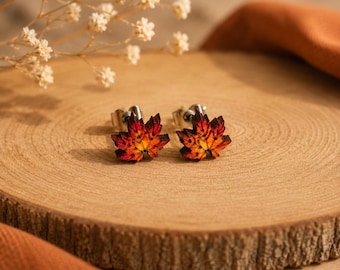Puede incluir: Un par de pendientes de botón con forma de hojas de arce, con un degradado de colores rojo, naranja y amarillo. Los pendientes tienen una textura detallada de nervaduras y están sujetos a postes de metal plateado con cierres redondos.