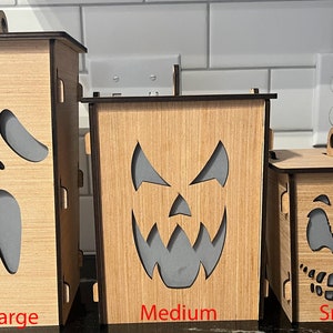 May include: Three wooden Halloween lanterns with different sized faces. The largest lantern has a screaming ghost face, the medium lantern has a jack-o-lantern face, and the smallest lantern has a grinning face. The lanterns are labeled "Large", "Medium", and "Small".